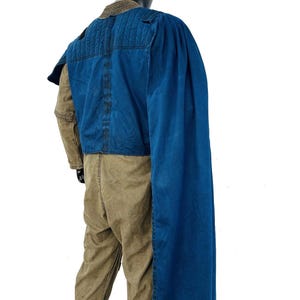 Back view of a Gar Saxon Mandalorian costume on a model, highlighting the long blue cape and the rear construction of the flak vest. Shows the full-length drape of the fabric and the tan flight suit pants, set against a professional white background.