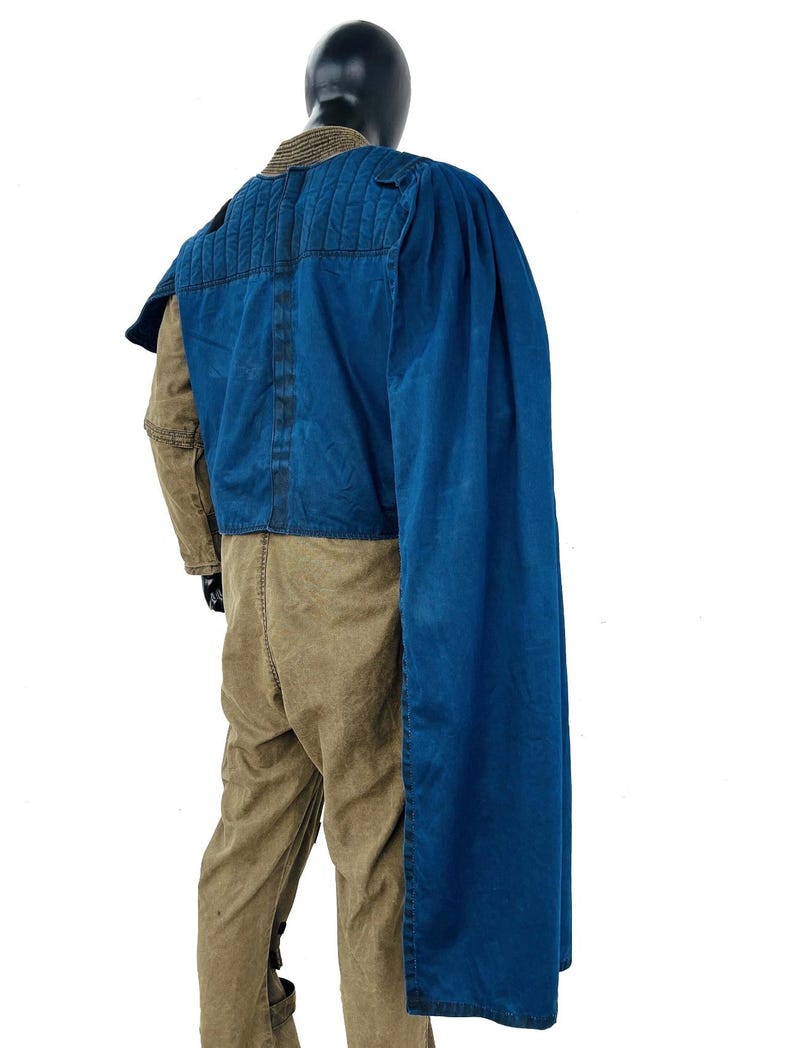 Back view of a Gar Saxon Mandalorian costume on a model, highlighting the long blue cape and the rear construction of the flak vest. Shows the full-length drape of the fabric and the tan flight suit pants, set against a professional white background.