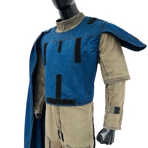 Front view of a male model wearing a Gar Saxon Mandalorian cosplay costume. Features a tan and grey flight suit, a blue tactical flak vest, and a matching long blue cape. Professional sci-fi costume displayed on a pure white background.