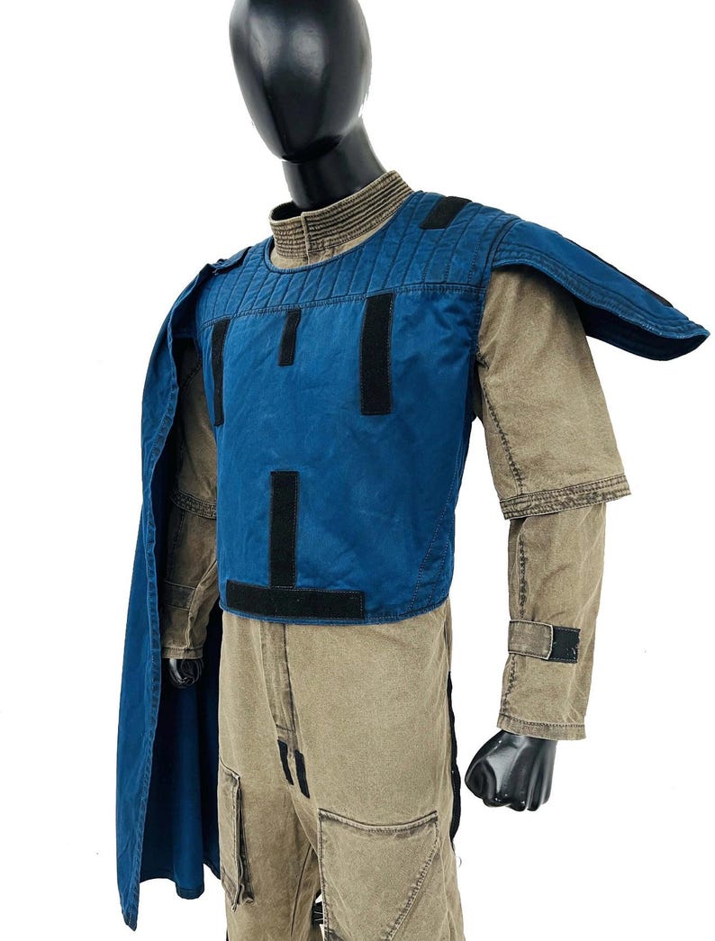 Front view of a male model wearing a Gar Saxon Mandalorian cosplay costume. Features a tan and grey flight suit, a blue tactical flak vest, and a matching long blue cape. Professional sci-fi costume displayed on a pure white background.