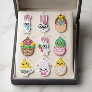 Pode incluir: Uma coleção de brincos bordados com tema de Páscoa. Os desenhos incluem coelhos, pintinhos, um cordeiro e um ovo com um dinossauro. Os brincos são exibidos em uma caixa de joias cinza com interior branco.