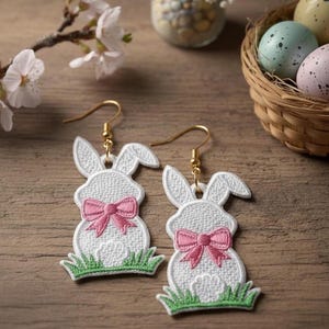 May include: Embroidered white bunny earrings with pink bows and green grass detail, featuring gold-coloured hooks. The image also shows a basket of speckled Easter eggs and a sprig of white flowers.