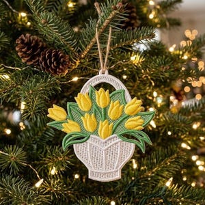 Könnte beinhalten: Gestickter Weihnachtsschmuck mit einem weißen Korb voller gelber Tulpen und grünen Blättern. Der Schmuck hängt an einer Schnur aus Naturfaser vor dem Hintergrund eines geschmückten Weihnachtsbaums mit Tannenzapfen und Lichtern.