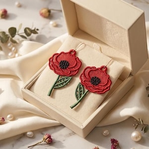 Könnte beinhalten: Ein Paar Ohrhänger mit gestickten roten Mohnblumen mit schwarzen Zentren und grünen Stielen. Die Ohrringe werden in einer offenen, cremefarbenen, rechteckigen Schmuckschatulle präsentiert, die mit weichem Stoff ausgekleidet ist und detaillierte Stickereien zeigt.