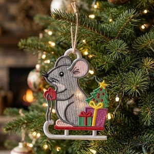 May include: A gray embroidered mouse ornament hanging by a jute string. The mouse sits on a red sled, holding a red mitten, with a green Christmas tree and two wrapped gift boxes in purple and green with yellow ribbon accents at its side.