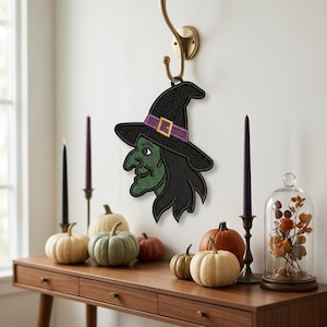 May include: A decorative embroidered wall hanging featuring a witch with green skin, a black hat with a purple band and yellow buckle, and black hair. The design is stitched with a textured pattern and hangs from a gold-colored metal hook.
