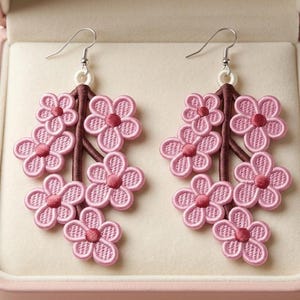 K&ouml;nnte beinhalten: Ein Paar rosa Blumenohrringe. Jeder Ohrring hat eine Ansammlung von gestickten rosa Blumen mit dunkelrosa Zentren, die an einem braunen Stiel befestigt sind. Die Ohrringe werden in einer rosa Schmuckschatulle pr&auml;sentiert.