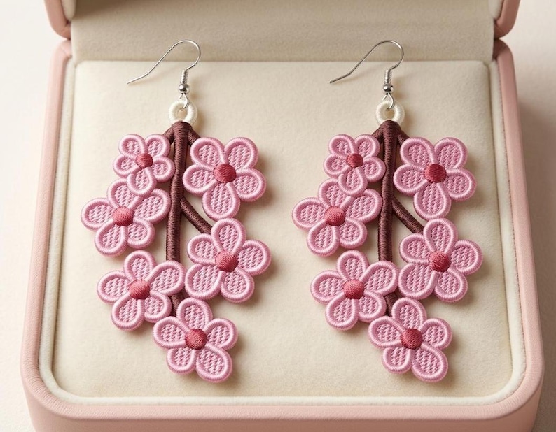 K&ouml;nnte beinhalten: Ein Paar rosa Blumenohrringe. Jeder Ohrring hat eine Ansammlung von gestickten rosa Blumen mit dunkelrosa Zentren, die an einem braunen Stiel befestigt sind. Die Ohrringe werden in einer rosa Schmuckschatulle pr&auml;sentiert.
