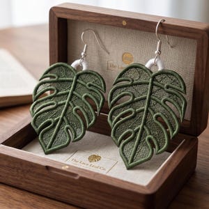 Puede incluir: Un par de pendientes verdes bordados con forma de hoja y ganchos plateados, expuestos en una caja de madera abierta. Los pendientes tienen forma de hojas de Monstera. La caja tiene un forro de tela beige y el texto "The Lace Leaf Co.".