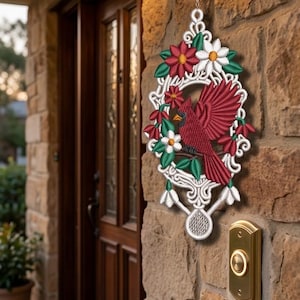May include: A decorative wall hanging featuring a red cardinal bird surrounded by red and white flowers and green leaves. The bird and floral design are set within a white, ornate frame. The hanging is displayed on a stone wall.