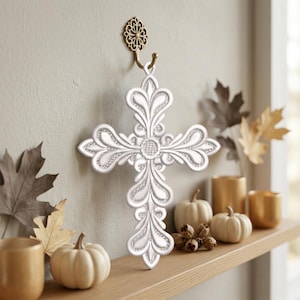 May include: A white decorative cross with intricate scrollwork and filigree patterns, suspended from a gold-toned metal wall hook. The cross features a central circular medallion and symmetrical, curved arms that terminate in rounded, petal-like shapes.