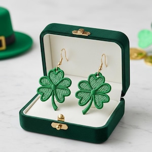 May include: A pair of green shamrock earrings with gold-colored hooks, displayed in an open, dark green velvet jewelry box. The earrings are embroidered with a detailed, textured design. A green leprechaun hat is visible in the background.