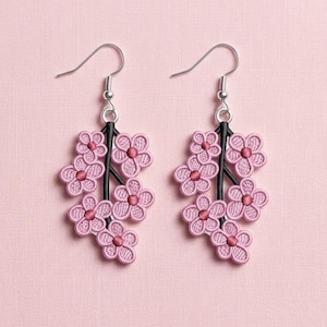 K&ouml;nnte beinhalten: Ein Paar rosa Blumenohrringe. Jeder Ohrring hat eine Ansammlung von gestickten rosa Blumen, die an einem schwarzen Stiel befestigt sind. Die Ohrringe haben silberfarbene Fischhaken-Ohrdr&auml;hte. Der Hintergrund ist einfarbig hellrosa.