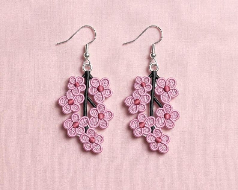 K&ouml;nnte beinhalten: Ein Paar rosa Blumenohrringe. Jeder Ohrring hat eine Ansammlung von gestickten rosa Blumen, die an einem schwarzen Stiel befestigt sind. Die Ohrringe haben silberfarbene Fischhaken-Ohrdr&auml;hte. Der Hintergrund ist einfarbig hellrosa.