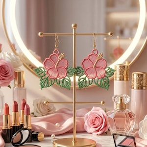 Puede incluir: Un par de pendientes colgantes con flores de hibisco rosas bordadas y hojas verdes. Los pendientes cuelgan de ganchos de metal dorado y se exhiben en un soporte de metal dorado con una base circular y barras horizontales.