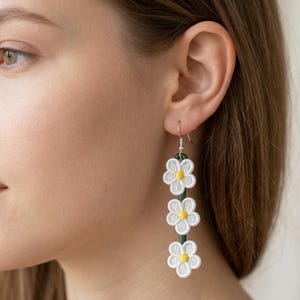 May include: White daisy earrings with yellow centers and green accents. The earrings are made of fabric and hang from a silver hook. The earrings are a floral design.