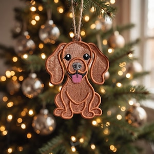 May include: A brown dog-shaped hanging ornament featuring detailed embroidery, large floppy ears, black eyes, a grey nose, and a pink tongue. The ornament is suspended by a thin, natural-colored jute twine loop for display on a tree branch.