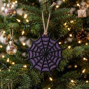 May include: A circular holiday ornament featuring a black spiderweb design layered over a textured purple background. The ornament is suspended by a natural jute twine loop, showcasing a detailed web pattern with radiating black spokes.