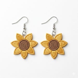 Puede incluir: Un par de pendientes de girasol. Cada pendiente presenta un girasol bordado amarillo con un centro marr&oacute;n y ganchos plateados. Los pendientes est&aacute;n sobre un fondo blanco.