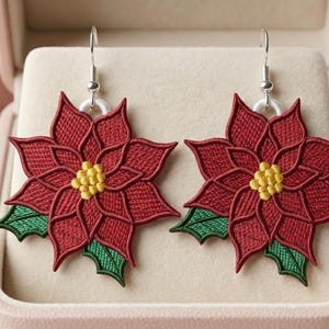 Puede incluir: Un par de pendientes de flor de Pascua. Cada pendiente presenta una flor roja bordada con centro amarillo y hojas verdes. Los pendientes se exhiben en una caja de joyería rosa.