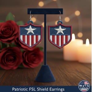 May include: Patriotic shield earrings with a star and stripes design. The earrings are displayed on a blue stand. The earrings have a blue background with a white star and red and white stripes. The text "Patriotic FSL Shield Earrings" is at the bottom.