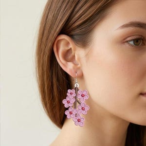 K&ouml;nnte beinhalten: Rosafarbene Blumenohrringe mit einem Astdesign. Die Ohrringe zeigen mehrere rosa Bl&uuml;ten mit roten Zentren, die an einem braunen Ast befestigt sind. Die Ohrringe h&auml;ngen an silbernen Haken.