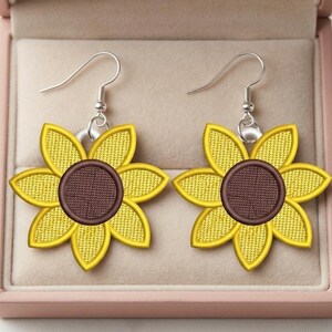 Puede incluir: Un par de pendientes de girasol con p&eacute;talos amarillos y centro marr&oacute;n. Los pendientes tienen ganchos plateados y se muestran en una caja de joyer&iacute;a rosa. Los pendientes est&aacute;n bordados con un dise&ntilde;o texturizado.