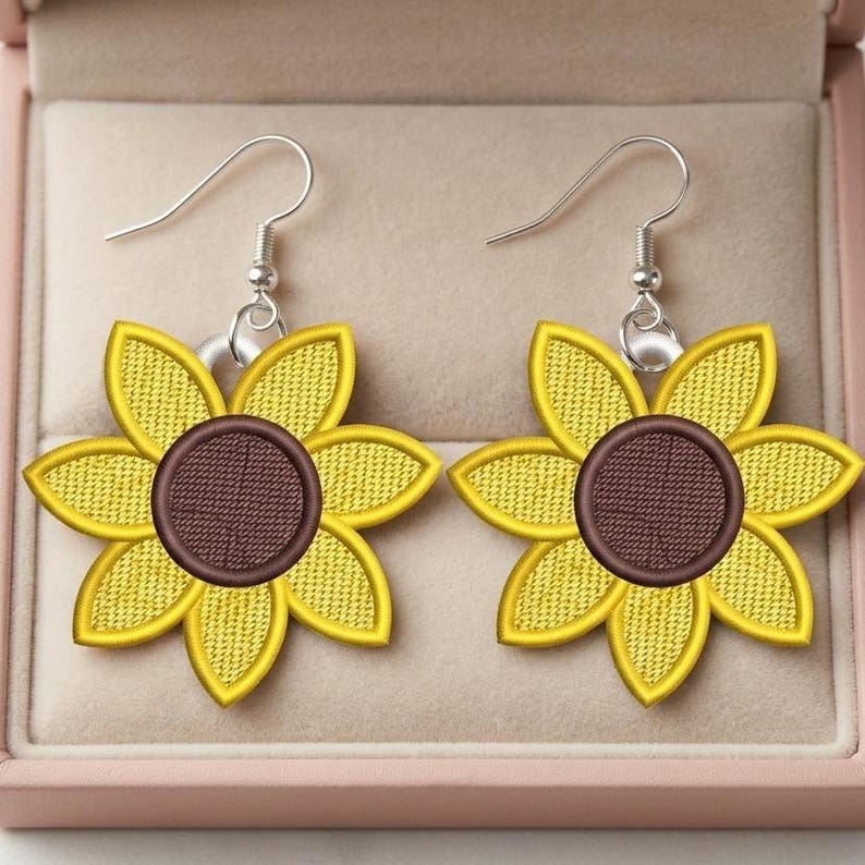 Puede incluir: Un par de pendientes de girasol con p&eacute;talos amarillos y centro marr&oacute;n. Los pendientes tienen ganchos plateados y se muestran en una caja de joyer&iacute;a rosa. Los pendientes est&aacute;n bordados con un dise&ntilde;o texturizado.