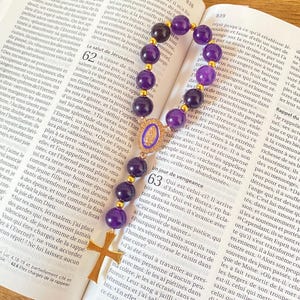 Può includere: Un rosario con perline viola e una croce e dettagli color oro, appoggiato su un libro aperto. Le perline sono di un viola intenso e la croce è dettagliata.