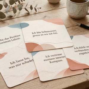 Könnte beinhalten: Eine Sammlung quadratischer Karten mit abgerundeten Ecken, die abstrakte Pastellmuster in Pfirsich, Blau und Beige aufweisen. Jede Karte hat deutschen Text. Die Karten liegen auf einer Holzoberfläche, zusammen mit einer Tasse, einem Stift und einer dekorativen Spule.