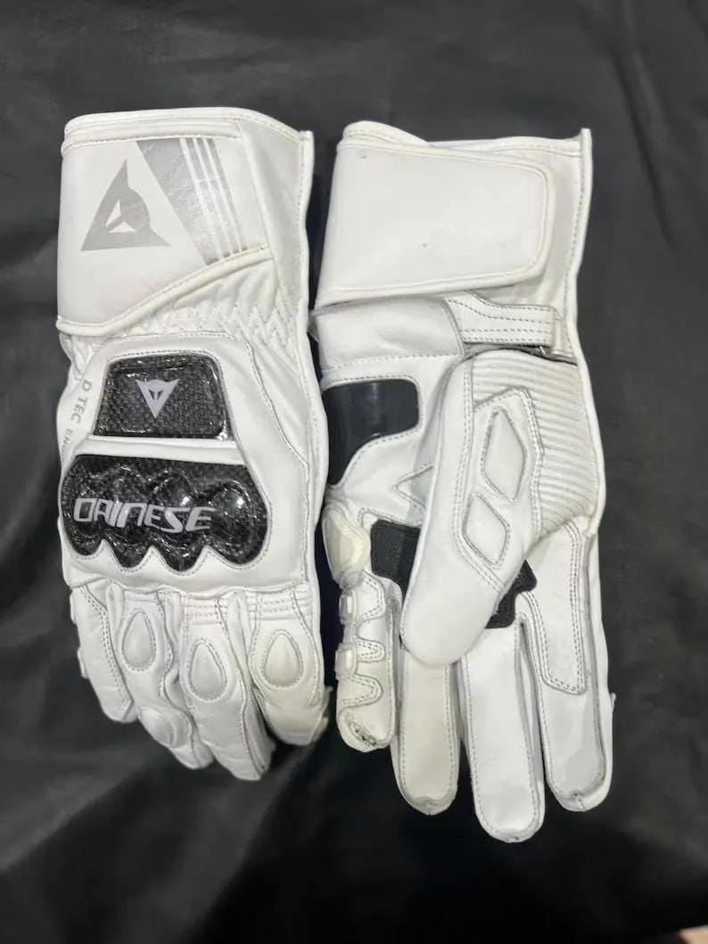Puede incluir: Par de guantes blancos de moto con detalles en negro. Los guantes muestran un logotipo y el nombre de la marca "Dainese". Tienen refuerzos en los nudillos y un cierre de mu&ntilde;eca seguro.