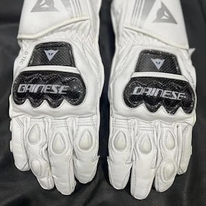 Puede incluir: Un par de guantes de moto blancos con detalles en negro y plata. Los guantes muestran la marca "DAINESE" y un logotipo en la mu&ntilde;eca y los nudillos. Tienen acolchado protector en los nudillos y los dedos.