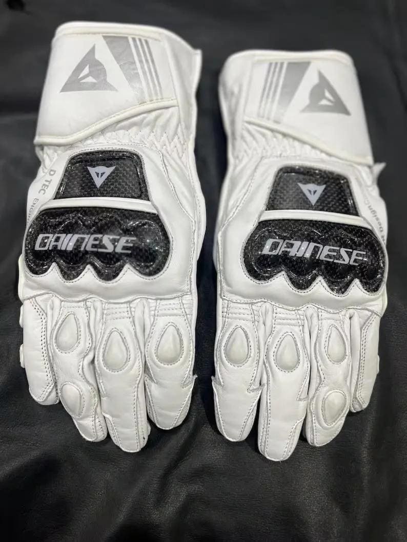 Puede incluir: Un par de guantes de moto blancos con detalles en negro y plata. Los guantes muestran la marca "DAINESE" y un logotipo en la mu&ntilde;eca y los nudillos. Tienen acolchado protector en los nudillos y los dedos.