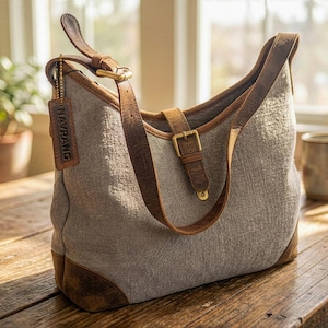 May include: A gray canvas shoulder bag with brown leather accents. The bag features a leather strap, a buckle closure, and a leather base. A tag with the text "NAVIRANG" hangs from the strap. The bag is sitting on a wooden table.