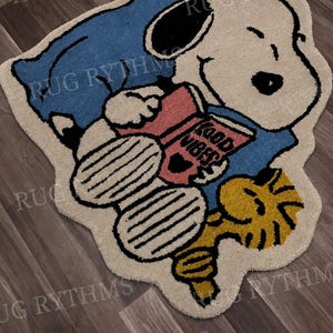 Peut inclure: Un tapis touffeté en forme de chien de dessin animé allongé sur un oreiller bleu, lisant un livre rose avec le texte "GOOD VIBES" sur la couverture. Un petit oiseau jaune est positionné près des pieds du chien. Le tapis est crème, bleu, rose et jaune.