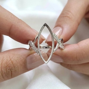 Könnte beinhalten: Ein silberfarbener Ring mit einer leeren, marquiseförmigen Fassung in der Mitte. Die Ringschiene ist an beiden Seiten in zwei sich kreuzende Abschnitte unterteilt, die jeweils mit kleinen, klaren, funkelnden Steinen besetzt sind.