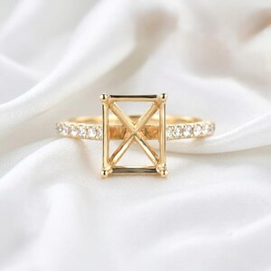 Könnte beinhalten: Eine goldene Ringfassung mit quadratischem Mittelteil und einem Band, das mit kleinen, klaren Edelsteinen besetzt ist. Der Ring ist vor einem weißen, seidigen Hintergrund platziert, der sein kompliziertes Design und seine Handwerkskunst hervorhebt. Dieser Ring ist perfekt für einen individuellen Verlobungsring.