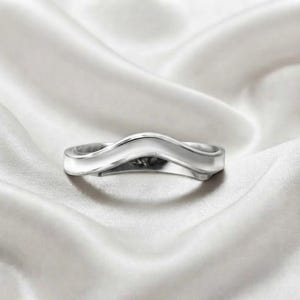 Könnte beinhalten: Ein polierter silberfarbener Ring mit einem glatten, geschwungenen V-förmigen Band. Die Metalloberfläche hat ein reflektierendes Finish und bildet eine klare, durchgehende Kontur, die in der Mitte abfällt und ein schlichtes Design zeigt.