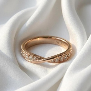 Puede incluir: Anillo de oro rosa con un diseño retorcido, incrustado con pequeñas piedras preciosas transparentes. El anillo tiene la inscripción "my love". El anillo se muestra sobre una tela blanca texturizada.