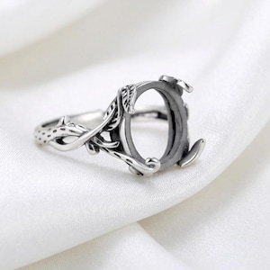 May include: A silver-toned ring with an oval setting, featuring a detailed leaf and vine design. The ring's band has a textured pattern, and the setting is designed to hold a gemstone. The ring is displayed on a white fabric background.