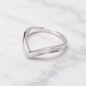 Könnte beinhalten: Ein silberner Ring mit einem einzigartigen V-förmigen Design. Der Ring hat eine glatte, polierte Oberfläche und eine moderne, minimalistische Ästhetik. Das Design des Rings weist ein spitzes Ende und ein gebogenes Band auf, was einen stilvollen und modernen Look erzeugt.