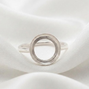 Könnte beinhalten: Ein silberner Ring mit einer kreisförmigen Fassung, die für einen Stein oder Cabochon ausgelegt ist. Der Ring hat eine glatte, polierte Ringschiene und eine leere, runde Fassung in der Mitte mit einem erhöhten Rand für die Schmuckherstellung.