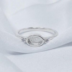 Könnte beinhalten: Ein silberfarbener Ring mit einer ovalen Fassung in der Mitte, die für einen Stein oder eine Einlage vorgesehen ist. Das Band ist auf beiden Seiten der zentralen Fassung mit einem kleinen, klaren, facettierten Stein verziert.