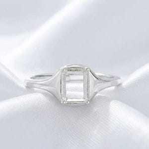 Könnte beinhalten: Ein silberfarbener Metallring mit einer rechteckigen, oben offenen Fassung, die zur Aufnahme eines Edelsteins dient. Der Ring hat eine glatte, polierte Ringschiene, die zur Mitte hin schmaler wird und eine klare Struktur für die Schmuckmontage bietet.