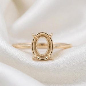 Puede incluir: Un engaste de anillo dorado con un centro de forma ovalada. El anillo tiene una banda delgada y cuatro garras para sujetar una piedra preciosa. Se encuentra sobre un fondo blanco y sedoso.