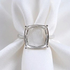 May include: A silver ring setting with a square cushion-shaped design. The ring has four prongs at each corner to hold a gemstone. The ring is displayed on a white fabric background.