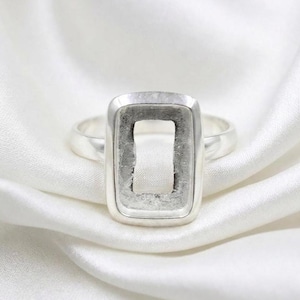 Könnte beinhalten: Ein silberner Ring mit rechteckiger Fassung. Der Ring hat einen polierten Ring und eine Lünettenfassung, die dazu bestimmt ist, einen Stein oder ein anderes dekoratives Element zu halten. Die Fassung ist leer und zeigt das Design.