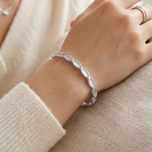 May include: A silver bracelet with a repeating oval pattern is shown on a wrist. The bracelet is made of a shiny metal and has a textured surface. The person is wearing a cream-coloured jumper.