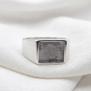 May include: A silver signet ring with a rectangular face. The face of the ring has a textured, gray appearance. The ring is set against a white background.