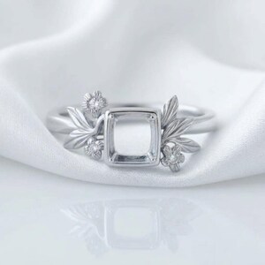 Könnte beinhalten: Ein silberfarbener Ring mit einer quadratischen Fassung in der Mitte, die für einen Stein vorgesehen ist. Die Ringschiene ist mit metallischen Blattmotiven und drei kleinen, runden, klaren Akzentsteinen im floralen Design verziert.